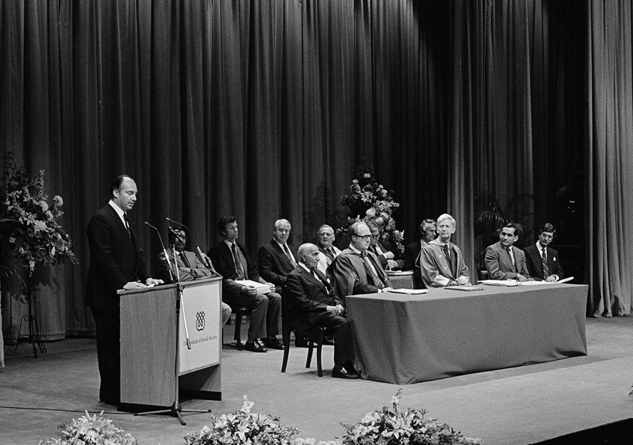 Following the presentation of the diplomas, Mawlana Hazar Imam addresses the new graduates at the University of London Institute of education. Seated at the front table with members of the Board of Governors of the Institute of Ismaili Studies (behind) are Mr. Dennis Lawson, Director of the Institute (left) and Mr. William Taylor, Principal of the University of London (right), July 1983.